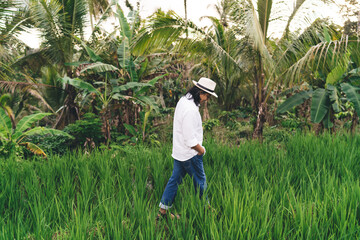Naklejka na ściany i meble Side view of Balinese man 40s in casual clothes walking around countryside during summer daytime in Indonesia, male farmer in hat visiting plantation with rice fields of paddy plants at Bali