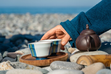 Close-up of a woman's hand taking a cup of tea from a wooden tray, there is a special teapot next to it. Tea ceremony on the pebble beach of the sea on a sunny day