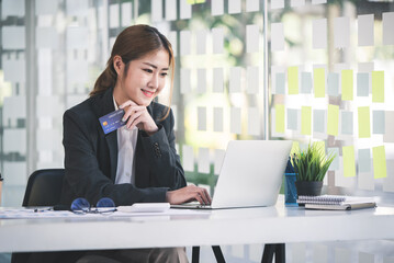 Asian woman holding a credit card and using a laptop computer Businesswoman working at modern office, online shopping, ecommerce, internet banking, spending money, credit card working concept.