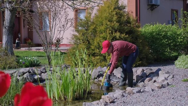 Adult Caucasian woman lets down a submersible water pump into an artificial garden fish pond to pump out water and clean the bottom from dirt and silt. Spring or Summer Pond Care