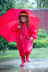 Happy funny child with red umbrella under  shower.Girl is wearing red  raincoat and enjoying rainfall. Kid playing on the nature outdoors.