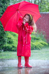 Happy funny child with red umbrella under  shower.Girl is wearing red  raincoat and enjoying rainfall. Kid playing on the nature outdoors.