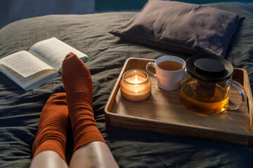 Soft photo of woman`s legs in the woolen socks on the bed with book and cup of tea and candle on...