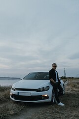 Handsome man in black T-shirt and white shirt with jeans and sneakers poses near modern white sport car and looks at the sunset outdoors. Fresh weekend out of the city near riverside. Dark tone photo
