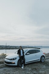Handsome man in black T-shirt and white shirt with jeans and sneakers poses near modern white sport car and looks at the sunset outdoors. Fresh weekend out of the city near riverside. Dark tone photo
