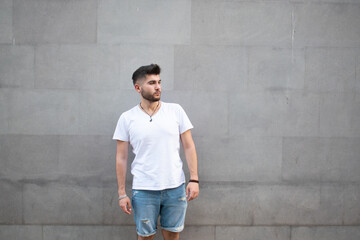 Bearded Armenian man with a white t-shirt and denim shorts posing on a wall background outdoors