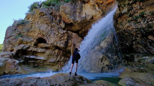 Woman is holding a mobile phone and taking a photo of a beautiful waterfall. Slow motion video. 