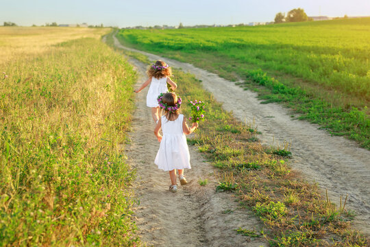 A girl in a white dress with a bouquet of clover flowers and a wreath in hair catches up with sister running along a sandy road into a field.