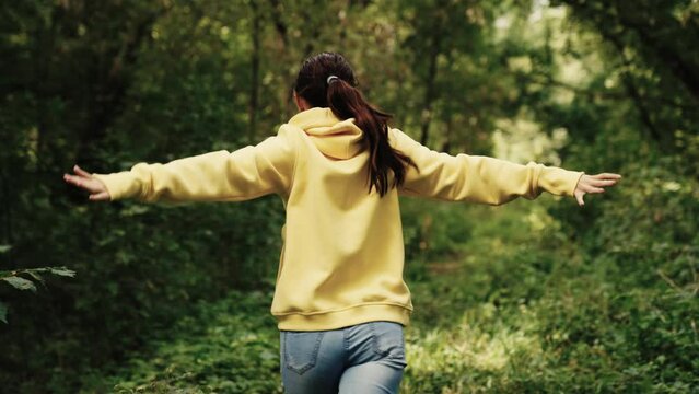 Young woman cheerfully runs in park. Happy person concept. Happy girl runs along the forest road waving her hands. Concept of joy, dreams, expression of emotions. Girl runs along path between trees.