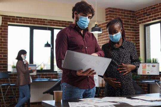 Multi Ethnic Diverse Office Workers Wearing Facemasks Against Coronavirus Analyzing Financial Data And Reviewing Client Portfolio. Expectant Mother Finance Advisor Looking Over Startup Project Ideas.