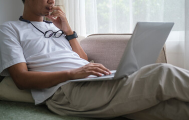 Young man in casual clothes surfing internet on laptop computer while sitting on couch at home.