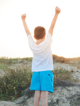 Preteen boy raises his arms in a sign of victory and strength looking towards the sun on a day at the beach dressed in a blue swimsuit and a white t-shirt.