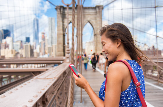 New York tourist woman using phone app walking on Brooklyn Bridge towards Manhattan city skyline. Young female professional multicultural lady, New York City, USA