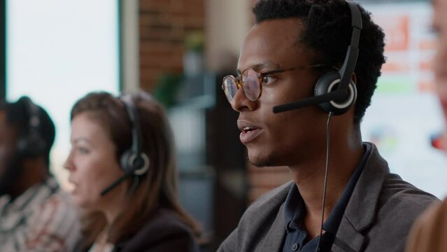 African american worker having conversation on headset with client, offering telemarketing assistance at call center. Man with headphones and microphone working at customer service. Close up.