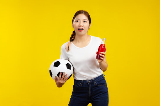 Surprised Asian woman over isolated yellow background holding a soccer ball and drinks, sport and people concept