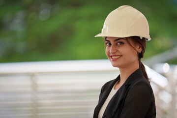 A beautiful woman engineer wearing white safety helmet look at camera with green bokeh background. Engineering and construction concept.