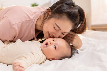 Obraz na płótnie Canvas New asian mom playing to adorable newborn baby on bed smiling and happiness at home.Mom talking with infant baby and kissing on baby cheek with love.Baby and Mother day concept