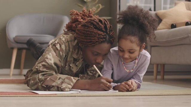 Slowmo of affectionate African American mother in military uniform and her pretty little daughter lying on stomachs on floor in living room and drawing in copybook