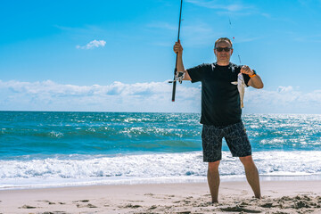 An elderly white caucasian man, 60 years old, holds a fishing rod and a freshly caught Whiting fish (pollock, Theragra chalcogramma) against the backdrop of the Atlantic Ocean in Florida. Copy space