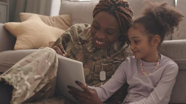 Slowmo of smiling young Black woman in military uniform and her 6 year old daughter drawing together sitting on floor by couch at cozy home