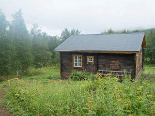 Fototapeta premium View of STF Njunjes Mountain cabin on a meadow, moody rainy day with thick fog. Wooden cottage lies on the banks of the Tarra river, at Padjelantaleden hiking trail. Lapland summer landscape