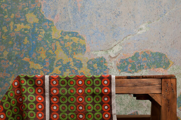 Fragment of old wooden table partially covered with tablecloth in geometric pattern in orange, green and brown colors. Multicolored, cracked wall in the background. Interior, rustic, retro style. 