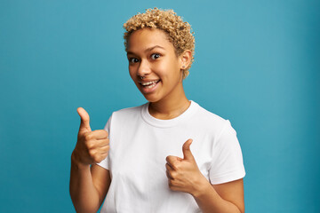 Happy surprised female with short blonde curls and noise piercing putting thumbs up over blue...