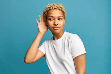 Good-looking curious dark-skinned female in white casual clothes putting hand over ear to hear better, listening to gossip or secret, having blonde curls, nose piercing and earrings, isolated on blue