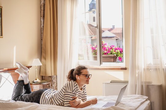 A young woman with glasses lying on the bed at home using a laptop, online work and education