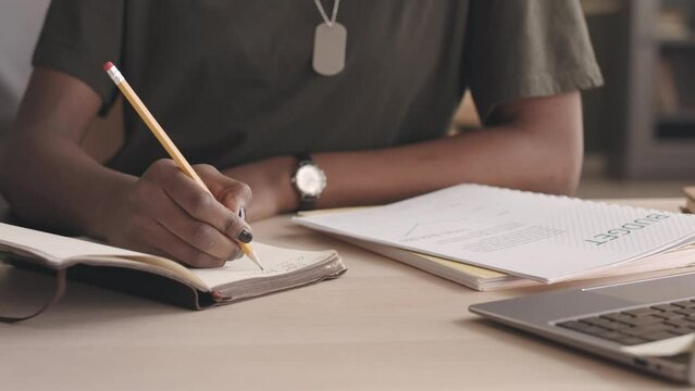 Cropped slowmo of unrecognizable Black woman in khaki t shirt taking notes in copybook while planning army budget working from home