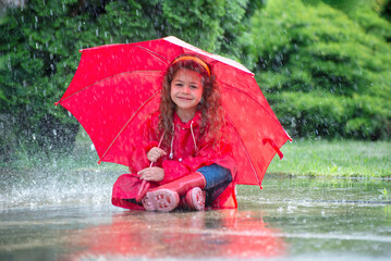 Happy funny child with red umbrella under  shower.Girl is wearing red  raincoat and enjoying rainfall. Kid playing on the nature outdoors.