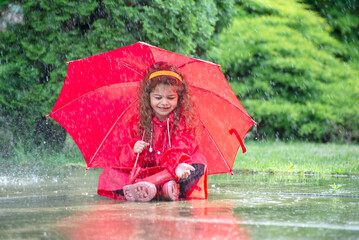Happy funny child with red umbrella under  shower.Girl is wearing red  raincoat and enjoying rainfall. Kid playing on the nature outdoors.