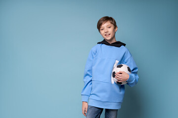 Smiling teen boy in sports blue clothes in a sweatshirt holding soccer ball posing in studio on a...