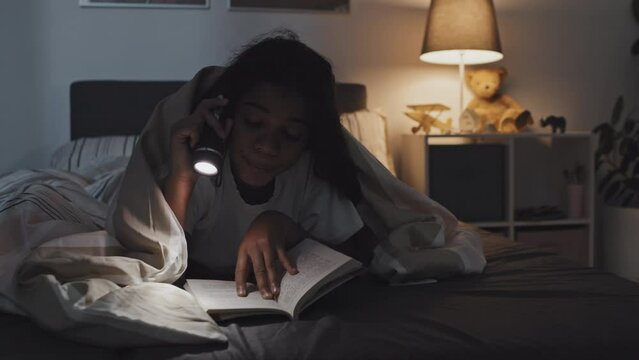 Chest-up of African American tween lying on stomach under blanket in bed, reading book using flashlight at night, yawning