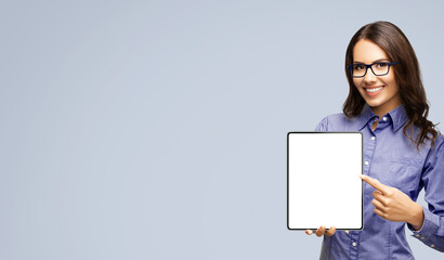 Naklejka premium Happy smiling businesswoman in eye glasses spectacles showing touchpad tablet pc, with white screen copy space text area for advertising or slogan sign, isolated over grey background. Business woman.