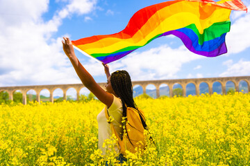 A black ethnic girl with braids holding the LGBT flag in a field of yellow flowers