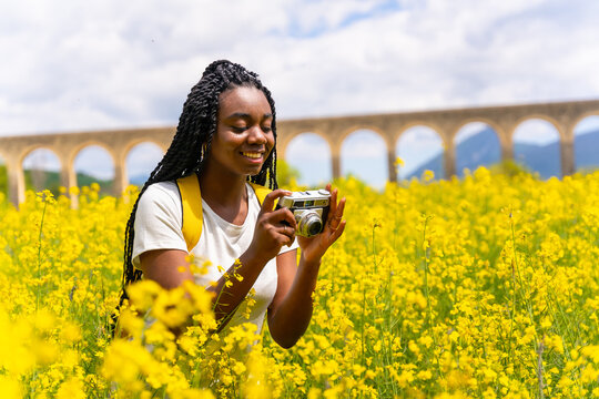 Taking photos of flowers with a vintage camera, a black ethnic girl with braids, a traveler, in a field of yellow flowers
