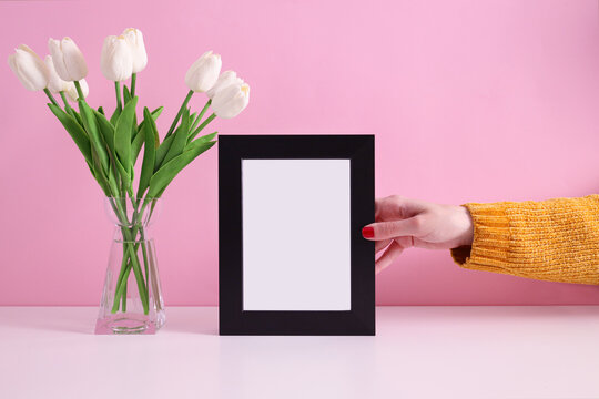 Young women holding photo frame with blank space for a text on a table with flowers bouquet, stylized image. Poster white frame mockup. Empty black frame mockup for presentation design. 