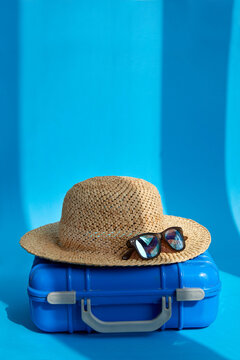 Plastic blue suitcase with straw hat and sunglasses placed on floor in bright studio for summer vacation