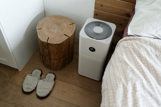 Modern technologies for health. An Electronic Air Purifier stands on the floor in the bedroom next to the bed with slippers and a wooden cabinet and cleans the air from dust.