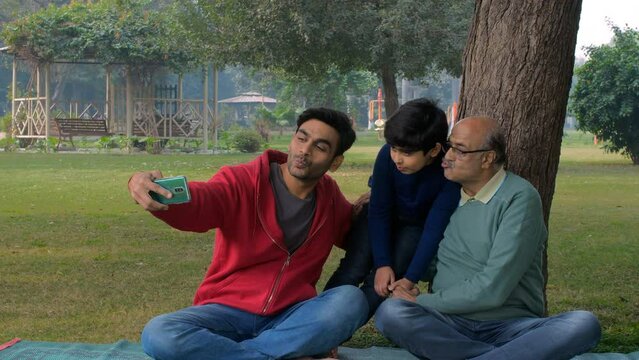 Indian family - Grandfather  grandson  and father clicking a selfie - picnic in a park  fun time. Three generations of male members posing for the camera in a public park - leisure time  bonding