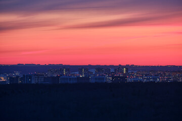 Night urban landscape with lights coming on in city houses. Multi-storey buildings after sunset with evening red sky