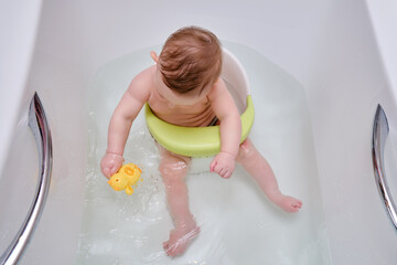 A happy infant baby bathes in water while sitting on a chair in a white tub. Toddler child plays...