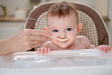 Mother feeding toddler baby from a spoon on a high chair for children, home kitchen background....
