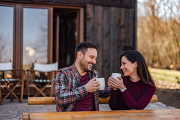 Smiling lovers enjoy the outdoor, sitting in front of their wooden cottage.