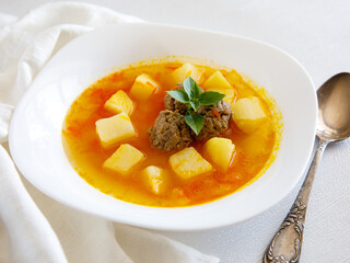 Delicious homemade meatball soup with vegetables potato, carrot and onion in white ceramic bowl on wooden table with spoon and napkin. Selective focus, top view