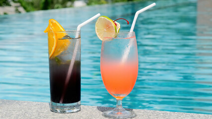 Vacation, summer, holiday, luxury resort concept. Two glasses with tropical cold cocktails with ice, plastic straw and slices of lime and orange near blue swimming pool in hot summer day