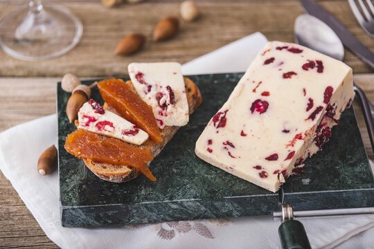 Wensleydale cheese with cranberries, red wine, honey, nuts, raisins on wooden cutting board. Black concrete background. Selective focus.