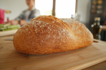 Whole loaf of bread on a wooden cutting board in the kitchen. Woman in background preparing breakfast sandwiches.
