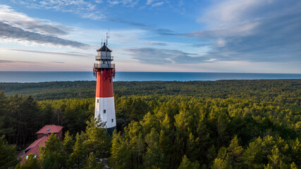 Aerial view of the lighthouse surrounded by the lush green forest. Sea in the distance. Blue sky and pastel clouds. Panoramic view.